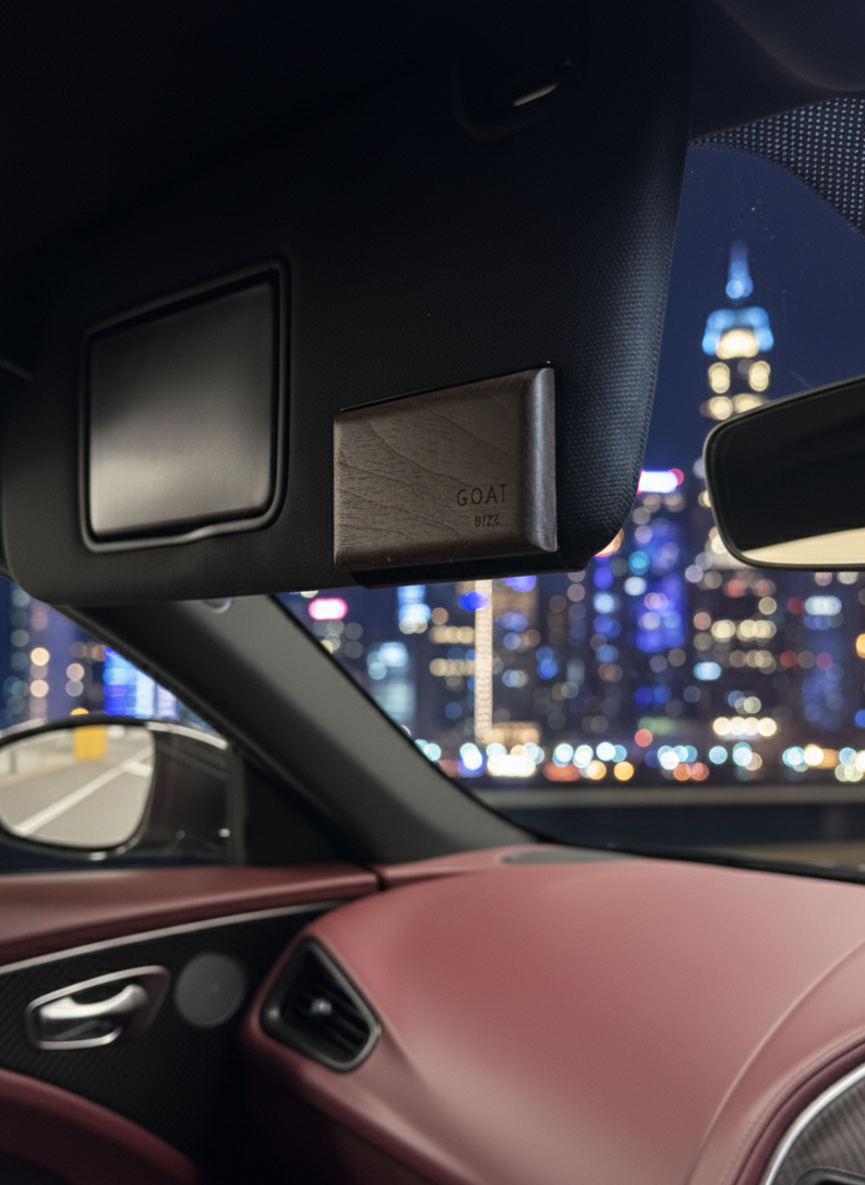 G.O.A.T BYZK Black Ice air freshener mounted on the sun visor of a luxury car with red leather interior, night city lights blurred in the background.